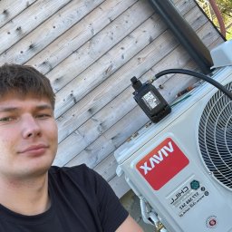 Chill Zone Mateusz Tomaszczuk - Instalator prezentuje zamontowaną klimatyzację XAVIV na drewnianej elewacji budynku. Widoczny czarny przewód i narzędzia serwisowe. Ujęcie z bliska.
