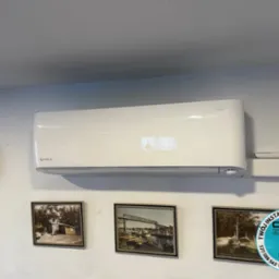 Biały klimatyzator Rotenso Mira na ścianie z zegarem i obrazami. Widoczny pilot. Logo firmy CHILL ZONE. Ujęcie z dołu.