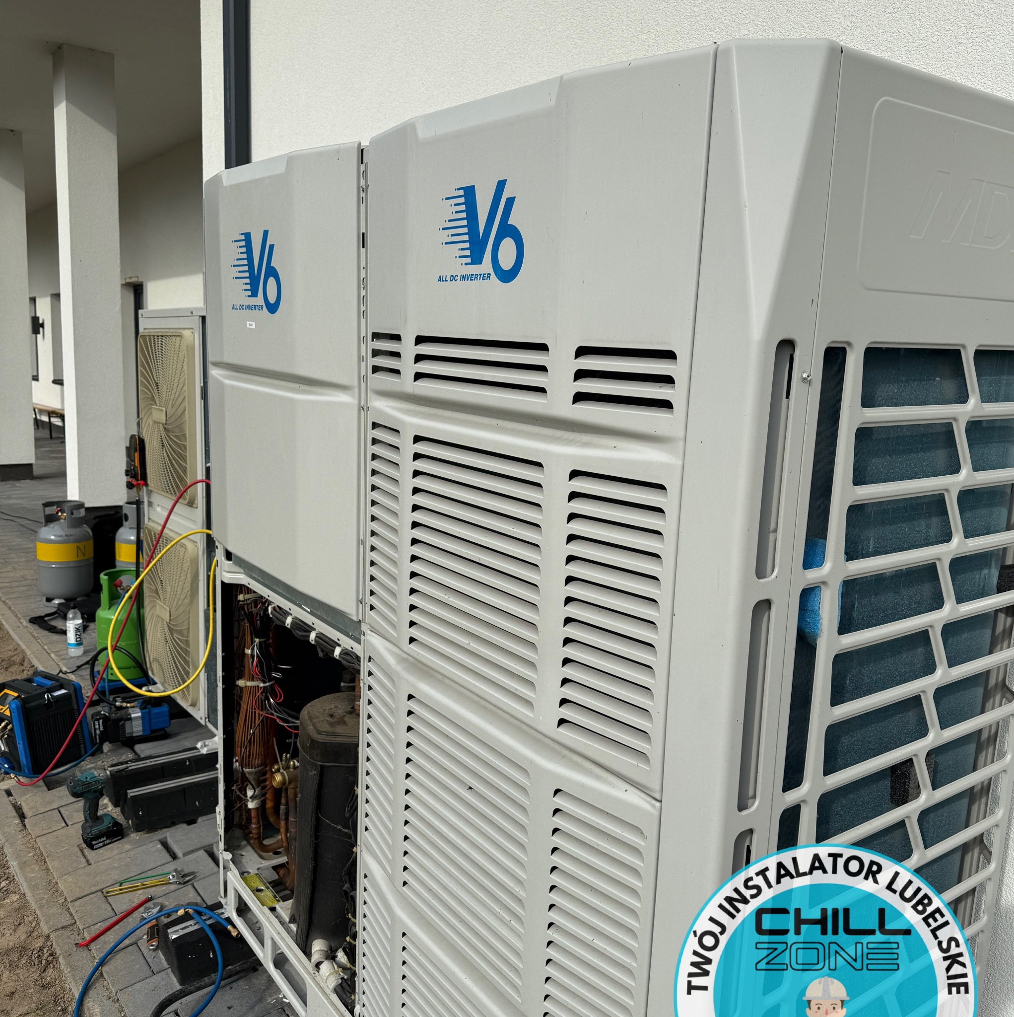 Dwa duże agregaty klimatyzacyjne V6 Inverter, jeden z otwartą obudową i widocznymi elementami instalacji, narzędzia serwisowe obok. Logo firmy na dole.