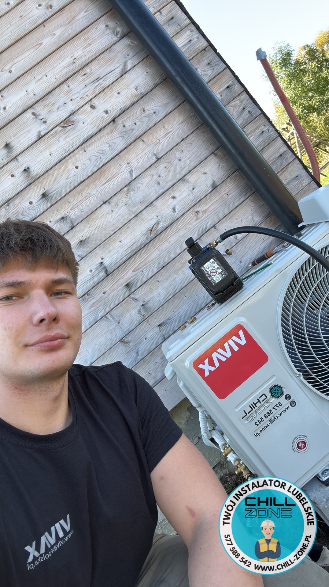 Instalator prezentuje zamontowaną klimatyzację XAVIV na drewnianej elewacji budynku. Widoczny czarny przewód i narzędzia serwisowe. Ujęcie z bliska.