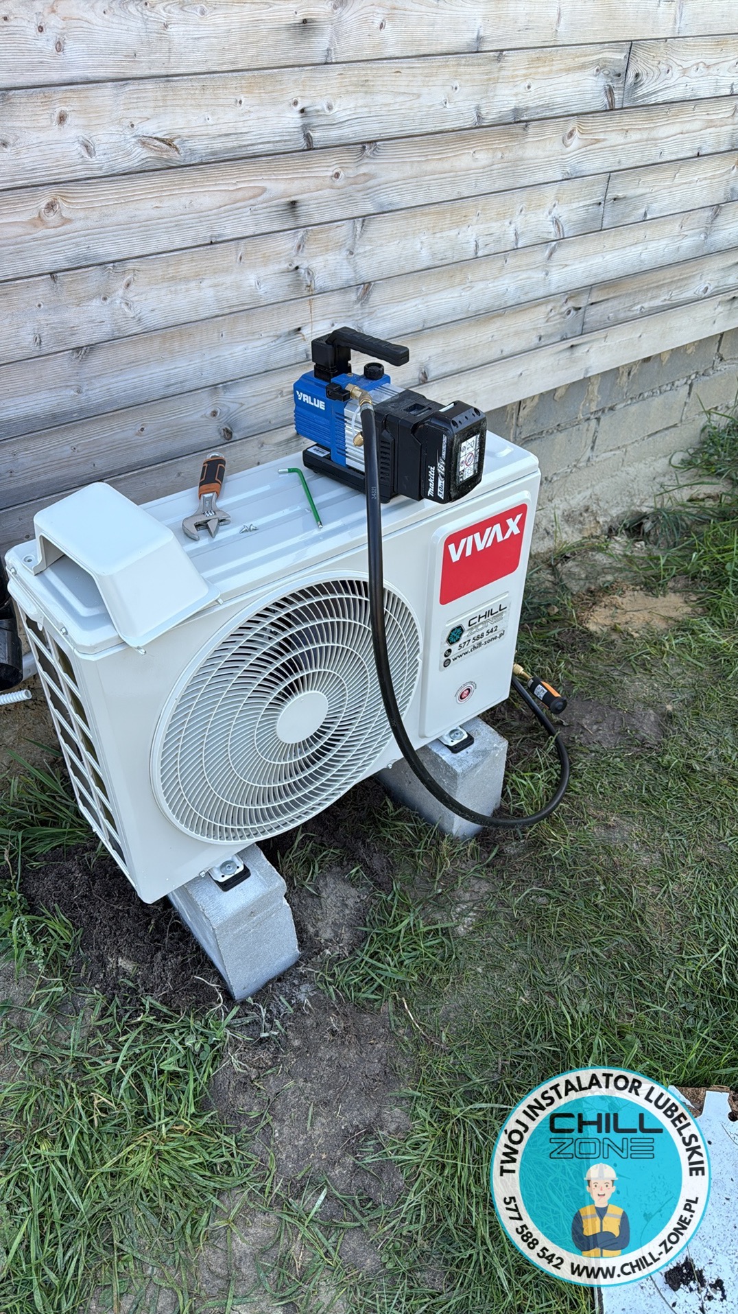 Biała jednostka zewnętrzna klimatyzacji VIVAX CHILL, stojąca na betonowych blokach, z podłączoną pompą próżniową VALUE i narzędziami, na tle drewnianej ściany i trawy.