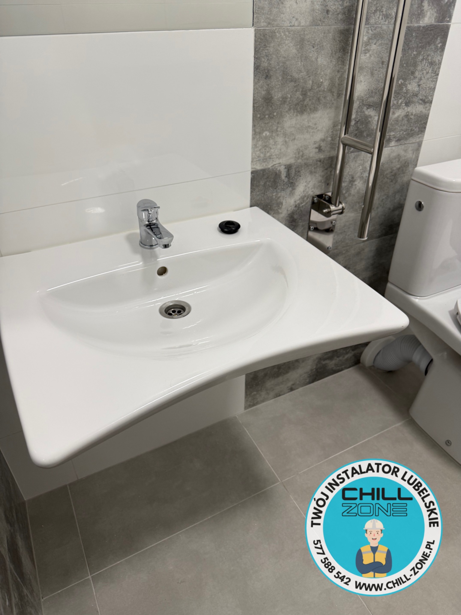 Biała umywalka z chromowaną baterią w nowoczesnej łazience z szaro-białymi płytkami. Widoczny fragment grzejnika drabinkowego i toalety. Na podłodze naklejka reklamowa.