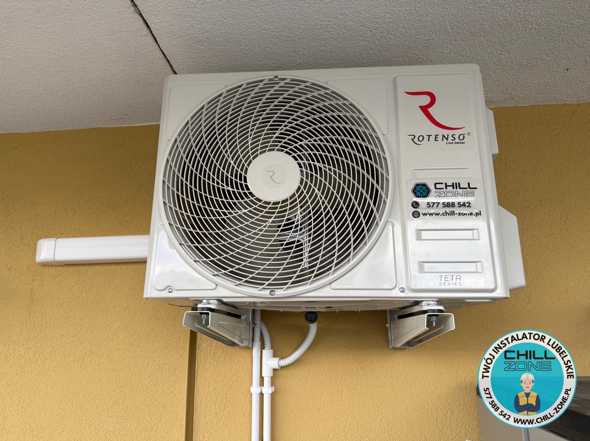 Biała jednostka zewnętrzna klimatyzacji Rotenso zamontowana na żółtej ścianie budynku. Widoczny fragment logo firmy instalacyjnej Chill Zone.