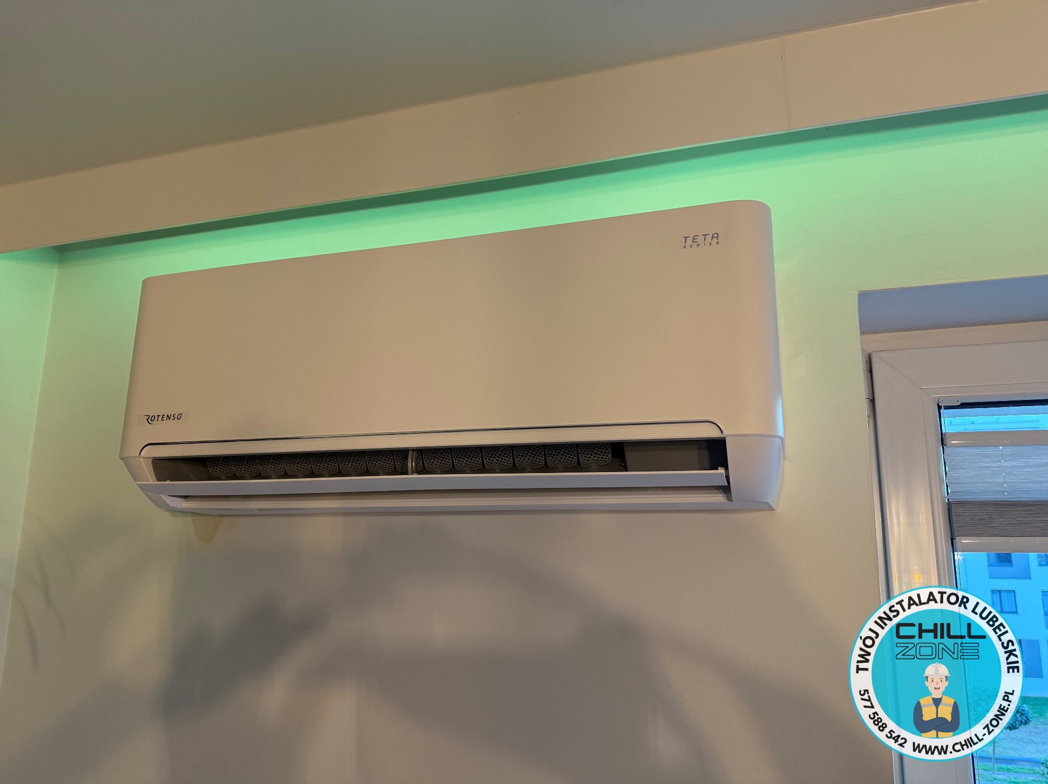 Biała klimatyzacja Rotenso Teta Series zamontowana na ścianie w pomieszczeniu z oknem. Widoczny logotyp firmy instalacyjnej.