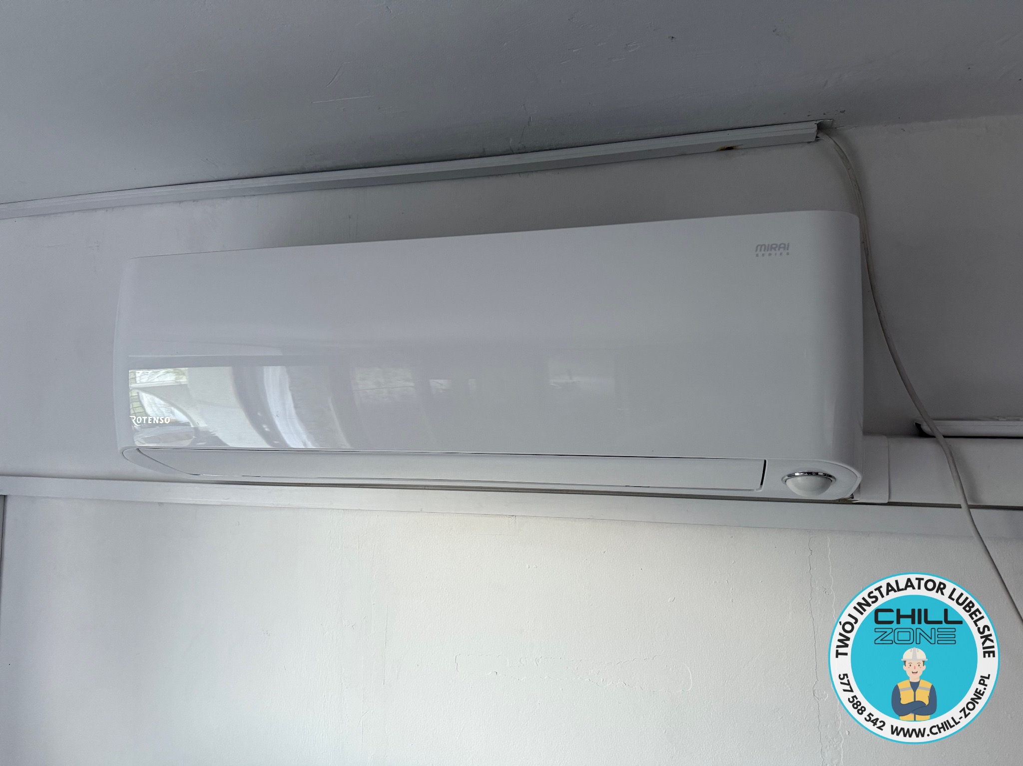 Biała klimatyzacja MIRAI zamontowana na ścianie z widocznym kablem zasilającym. Na ścianie widoczne logo firmy instalacyjnej Chill Zone Lubelskie.