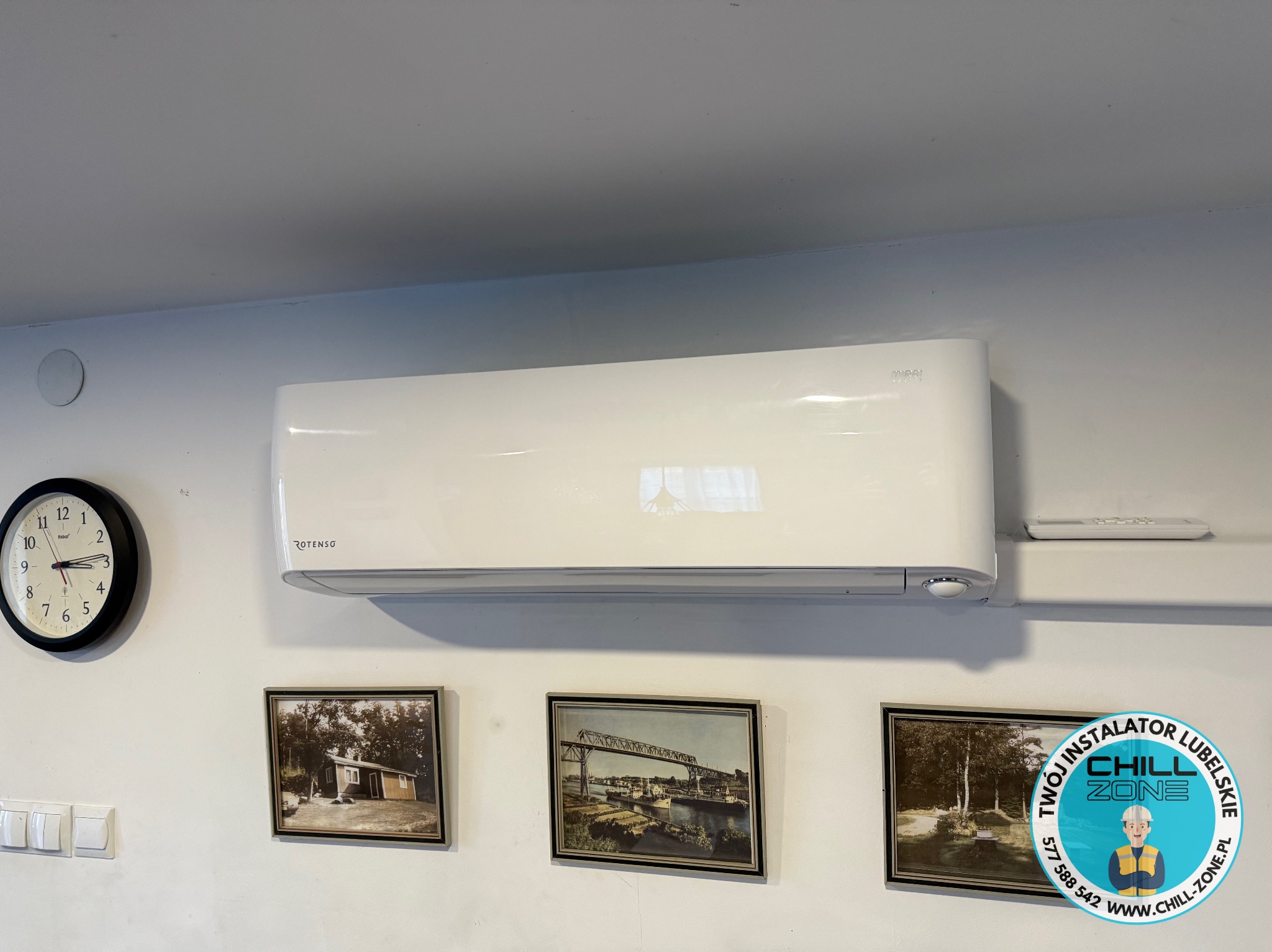 Biały klimatyzator Rotenso Mira na ścianie z zegarem i obrazami. Widoczny pilot. Logo firmy CHILL ZONE. Ujęcie z dołu.