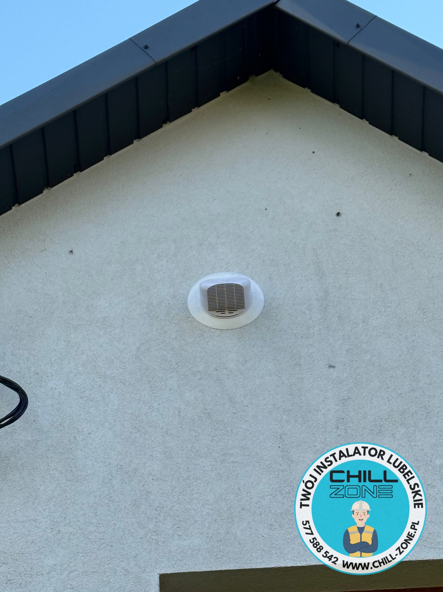 Biała kratka wentylacyjna na elewacji domu w Lublinie, zamontowana pod szczytem dachu. Widoczne logo firmy instalacyjnej.