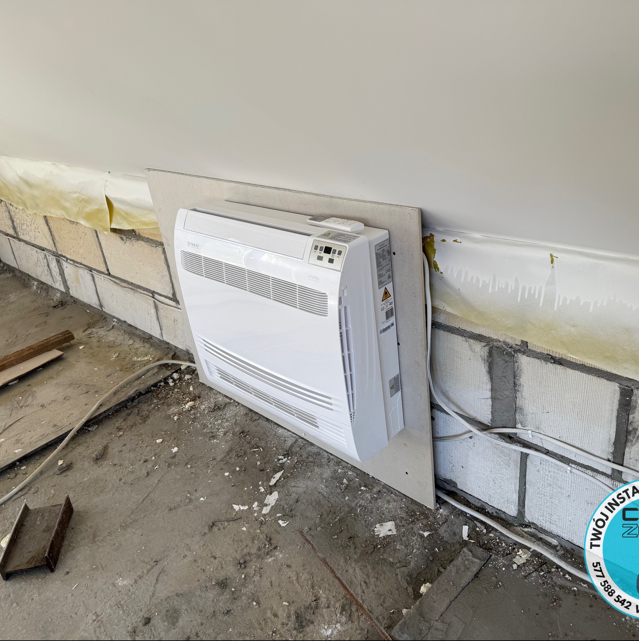 Biała klimatyzacja podłogowa Rotenso, zamontowana na płycie gipsowej przy ścianie z bloczków w surowym wnętrzu poddasza. Widoczne przewody i elementy montażowe.