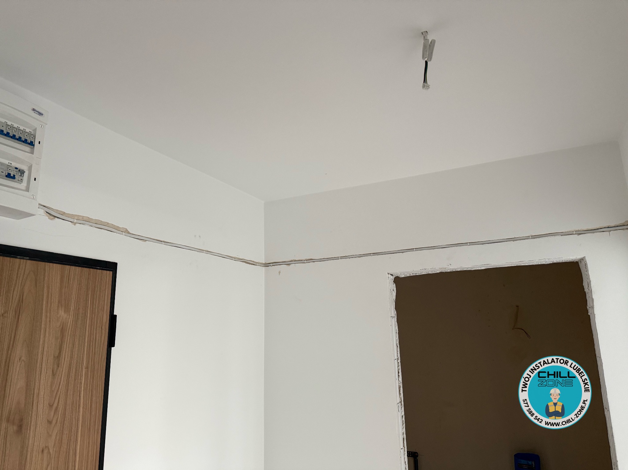 Białe wnętrze w trakcie prac instalacyjnych: widoczna skrzynka elektryczna, przewody pod sufitem i otwór w ścianie. Logo firmy w prawym dolnym rogu.