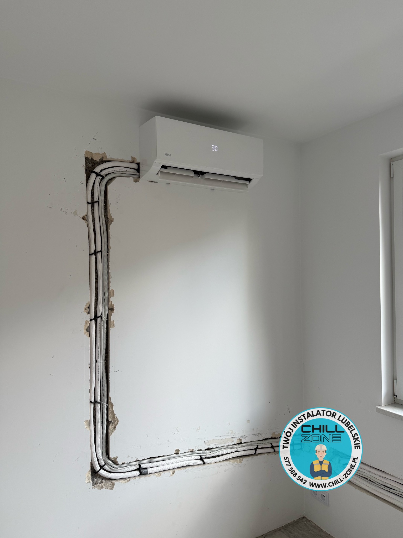 Biała klimatyzacja na ścianie z widocznymi przewodami w listwach, instalacja w trakcie montażu, ściana z ubytkami, logo firmy w rogu.
