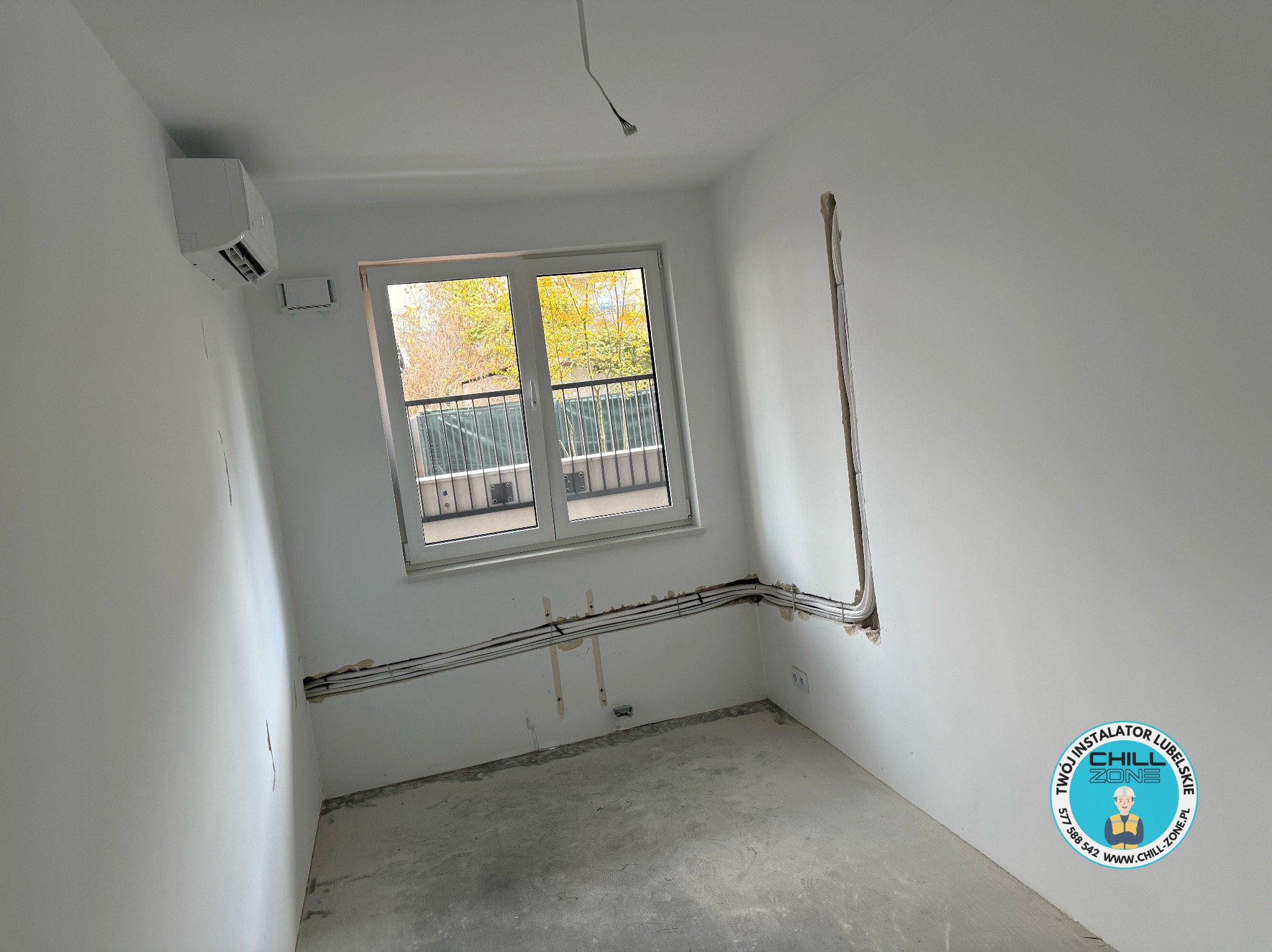 Białe wnętrze z zamontowaną klimatyzacją ścienną, widoczne przewody instalacyjne w ścianie, okno z widokiem na zieleń. Minimalistyczny design, surowy stan wykończenia.