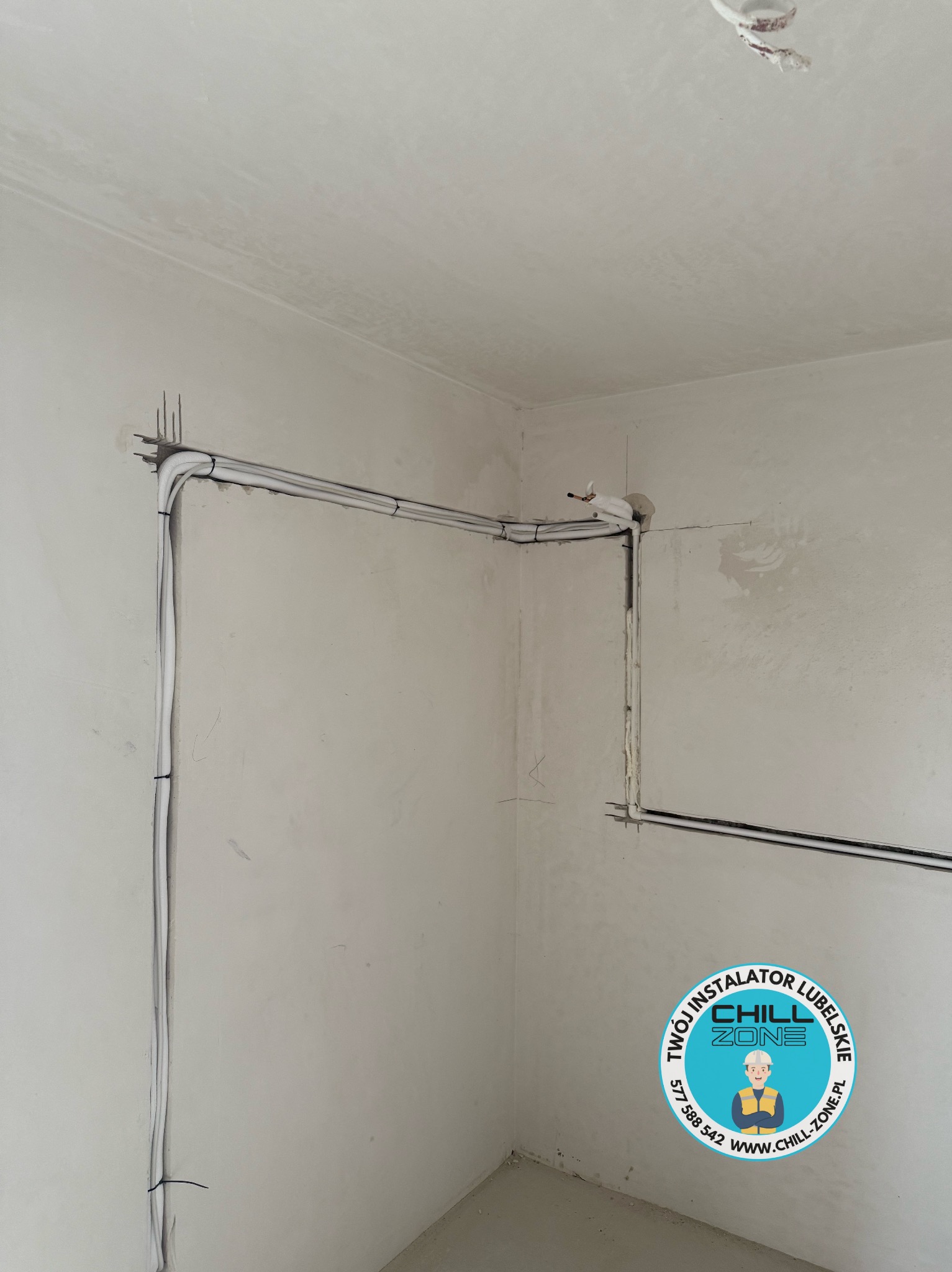 Surowy stan: białe rury instalacji klimatyzacyjnej poprowadzone wzdłuż ścian w narożniku pomieszczenia, zabezpieczone czarnymi opaskami. Widoczne ślady po kuciu w ścianie.