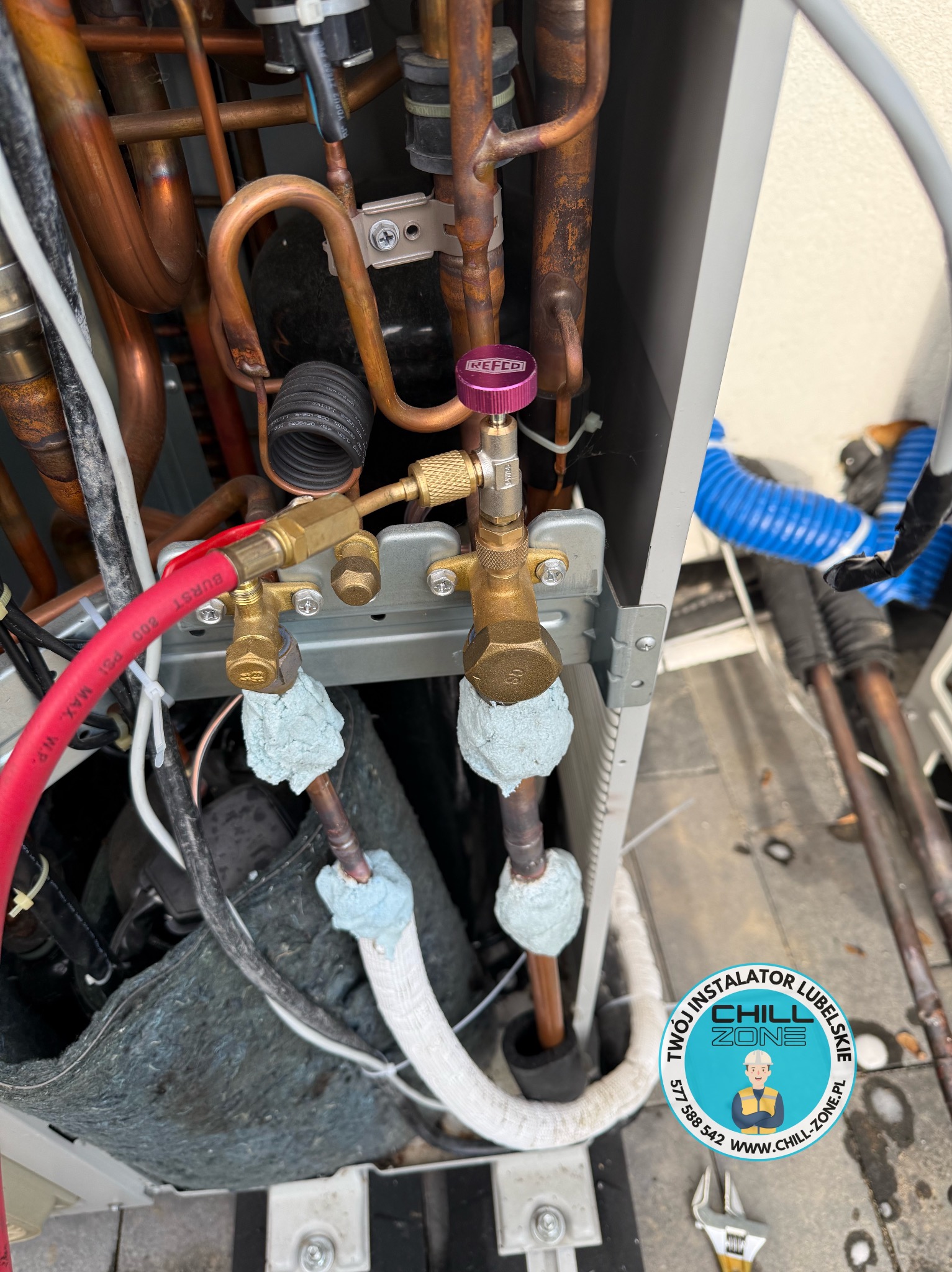 Wnętrze jednostki klimatyzacyjnej z miedzianymi rurami, zaworami REFCO i izolacją. Widoczny czerwony przewód i narzędzie monterskie. Logo firmy w prawym dolnym rogu.