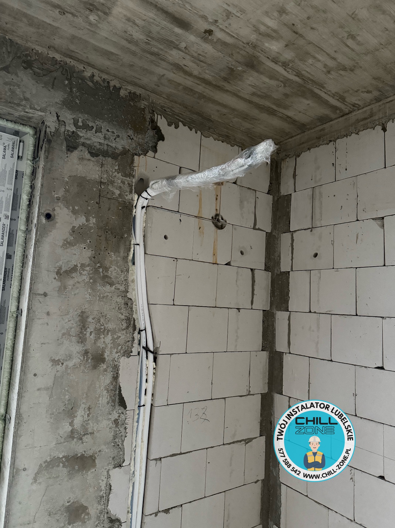 Surowy stan: białe rury instalacyjne prowadzone wzdłuż ściany z betonu komórkowego, częściowo owinięte folią ochronną. Widoczne otwory w ścianie i logo firmy.
