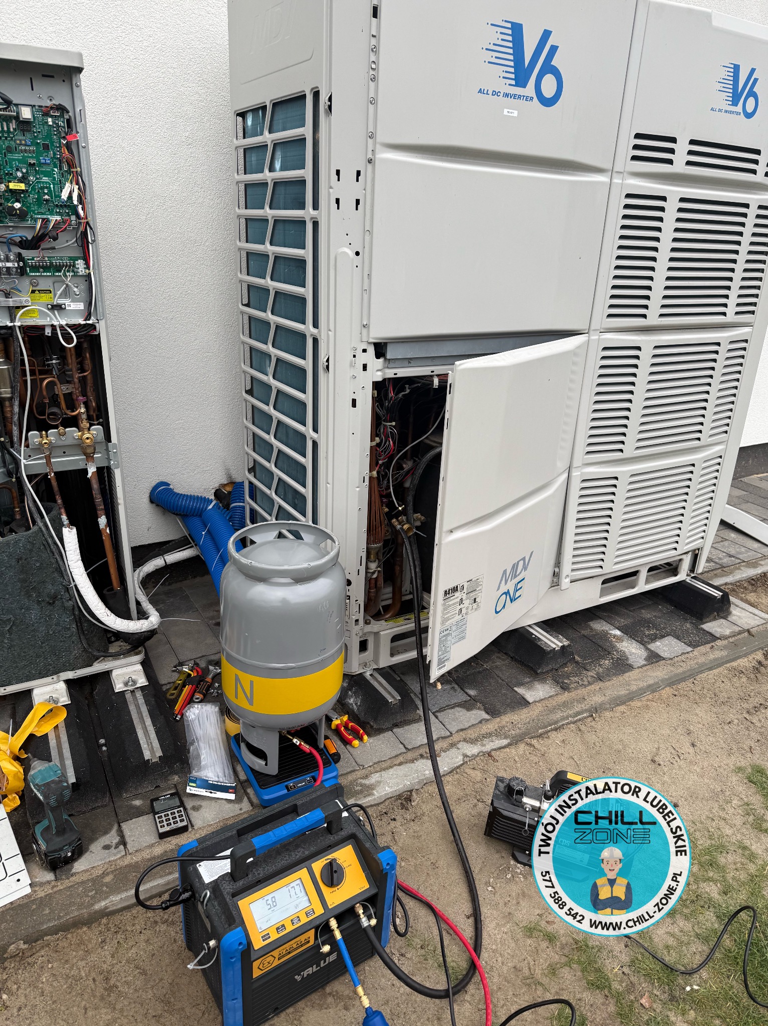 Serwis klimatyzacji MDV ONE V6: otwarte panele, widoczne komponenty, narzędzia serwisowe, butla z gazem. Prace serwisowe w toku, logo firmy w prawym dolnym rogu.