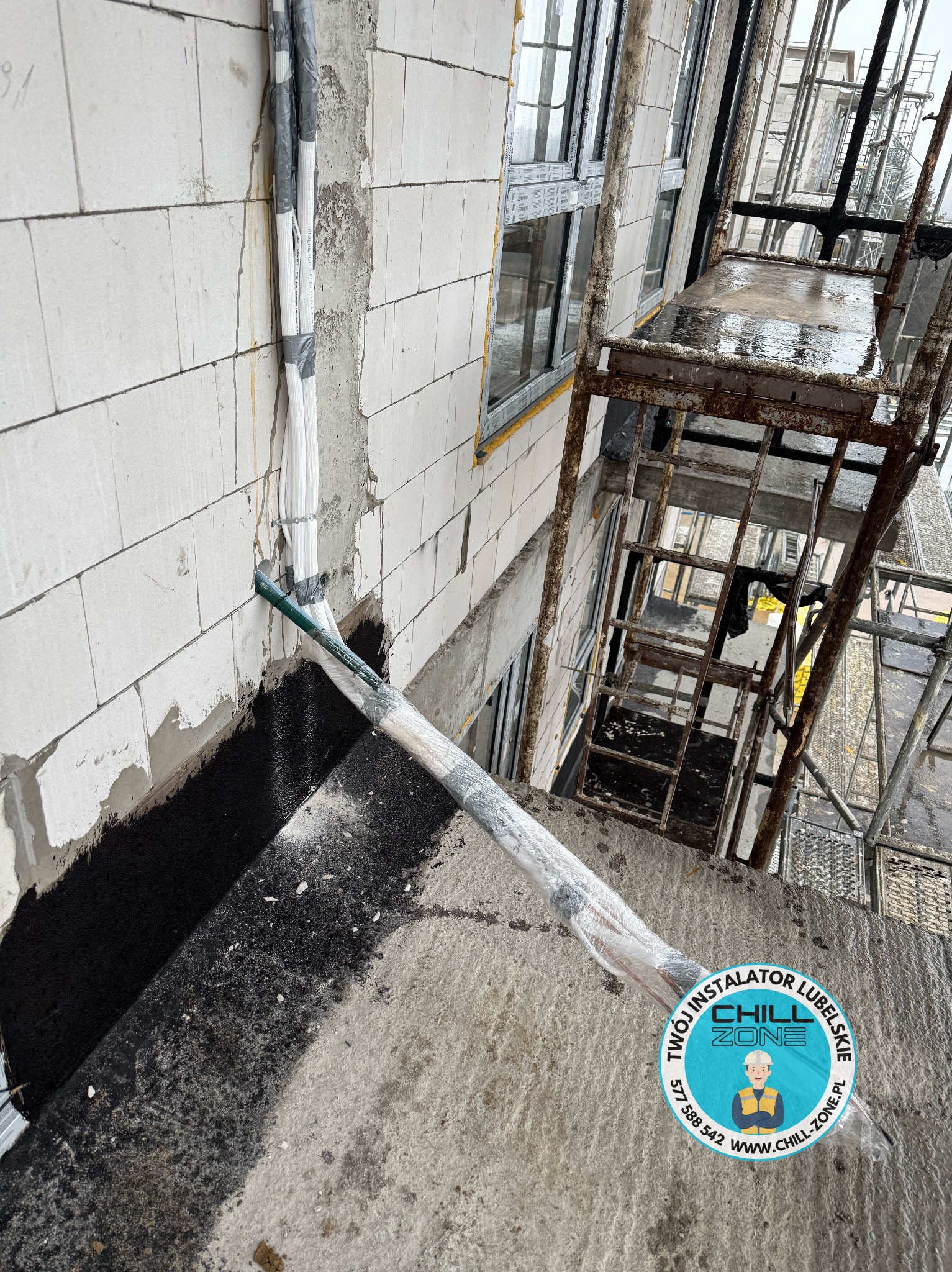 Montaż przewodów wentylacyjnych na ścianie z betonu komórkowego, zabezpieczonych folią. W tle rusztowanie i okna. Widoczny logotyp firmy.