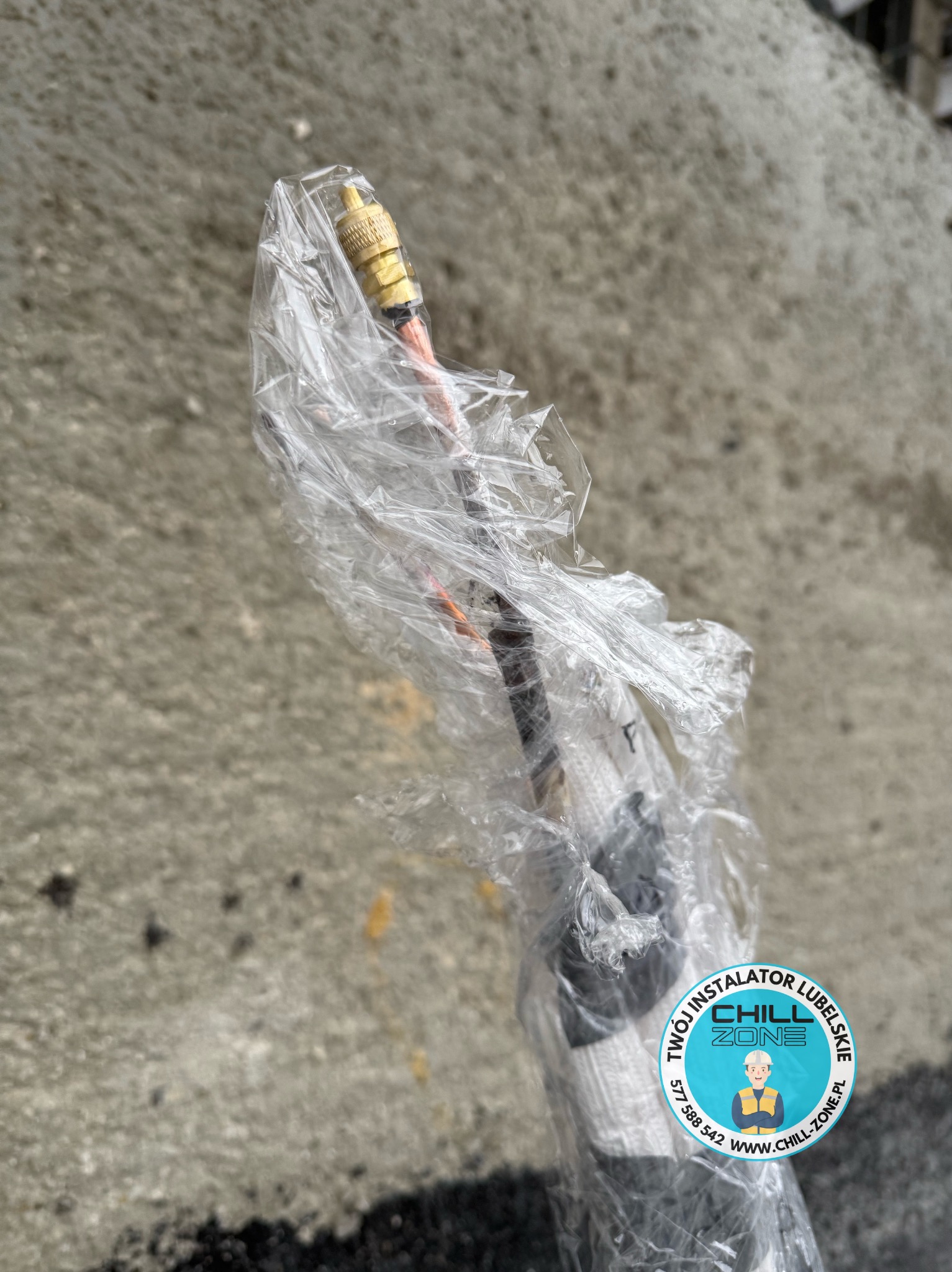 Kabel antenowy z wtyczką F, owinięty folią ochronną, z naklejką firmy instalacyjnej Chill Zone Lubelskie. Widok z bliska na tle betonowej powierzchni.