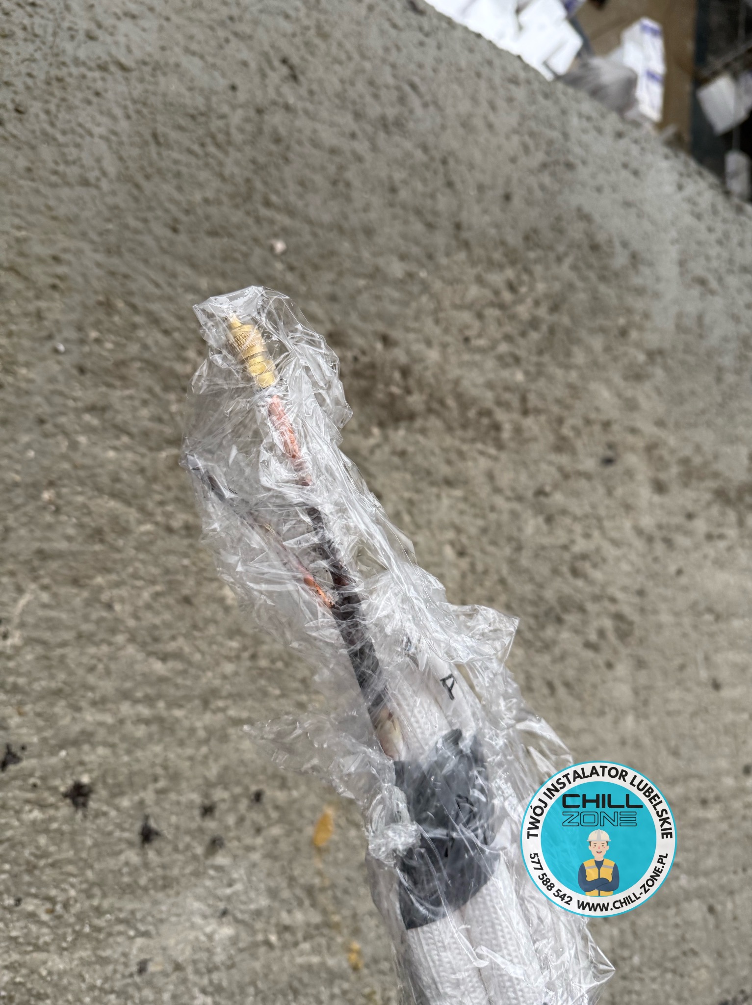 Kabel w izolacji termicznej, zabezpieczony folią, z widocznym logo firmy instalacyjnej Chill Zone Lubelskie. Szary beton w tle. Detal instalacji.