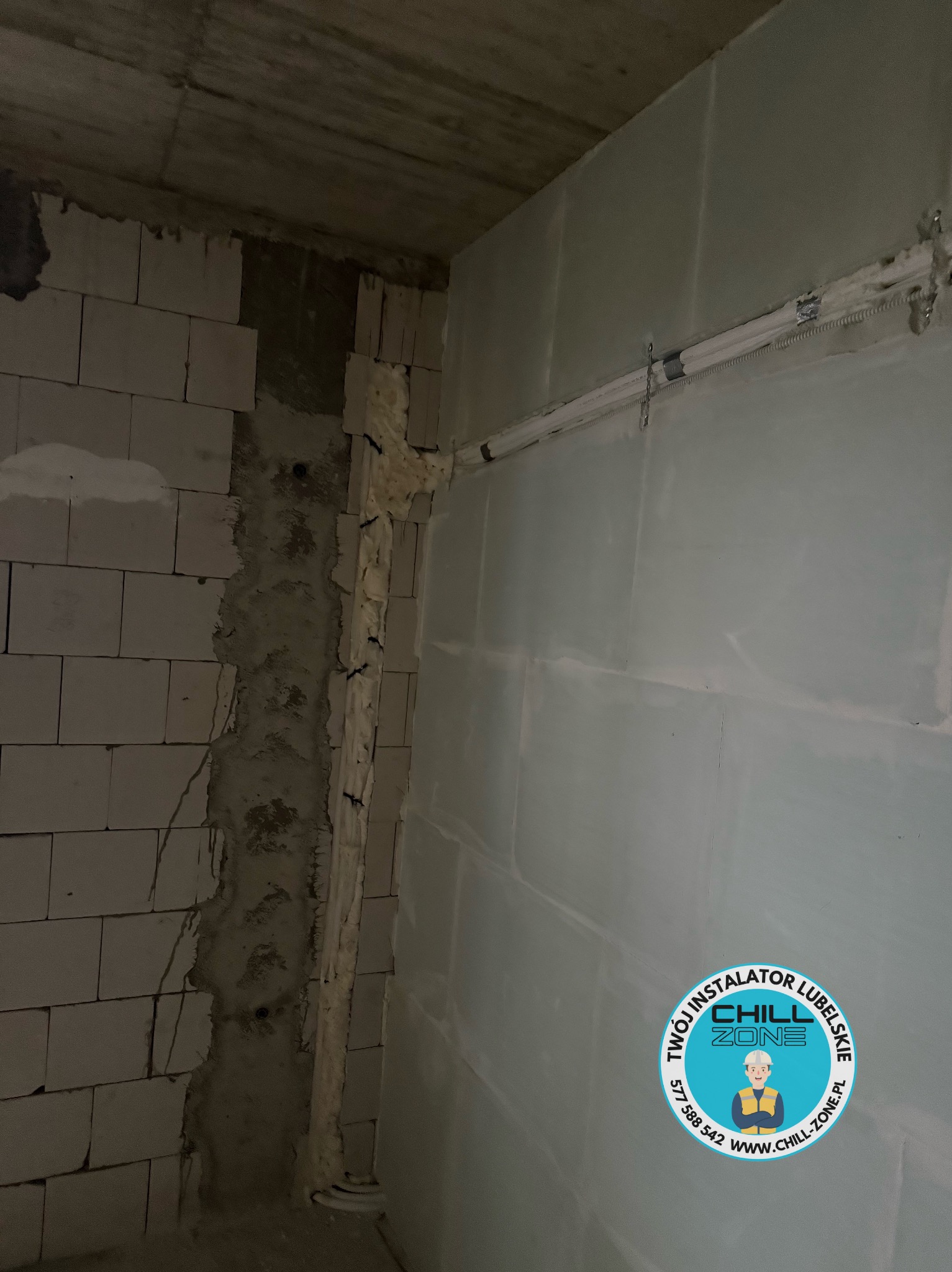 Montaż instalacji w surowym wnętrzu: widoczne przewody w ścianie, częściowo zakryte pianką montażową i płyty gipsowe. Logo firmy w prawym dolnym rogu.