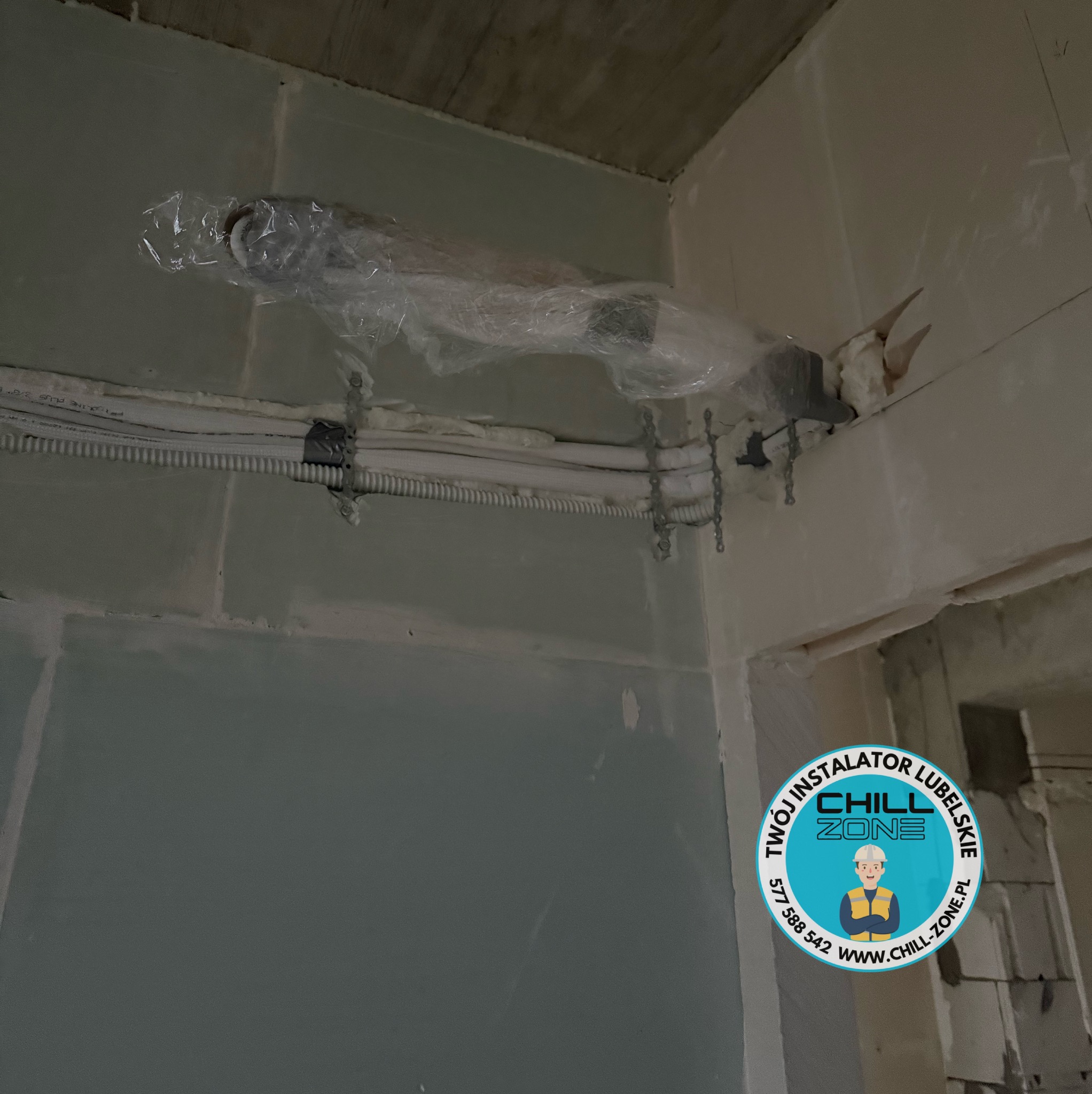 Montaż instalacji wentylacyjnej w surowym wnętrzu. Rury zabezpieczone folią, przymocowane do sufitu. Widoczne przewody i pianka montażowa. Logo firmy w rogu.