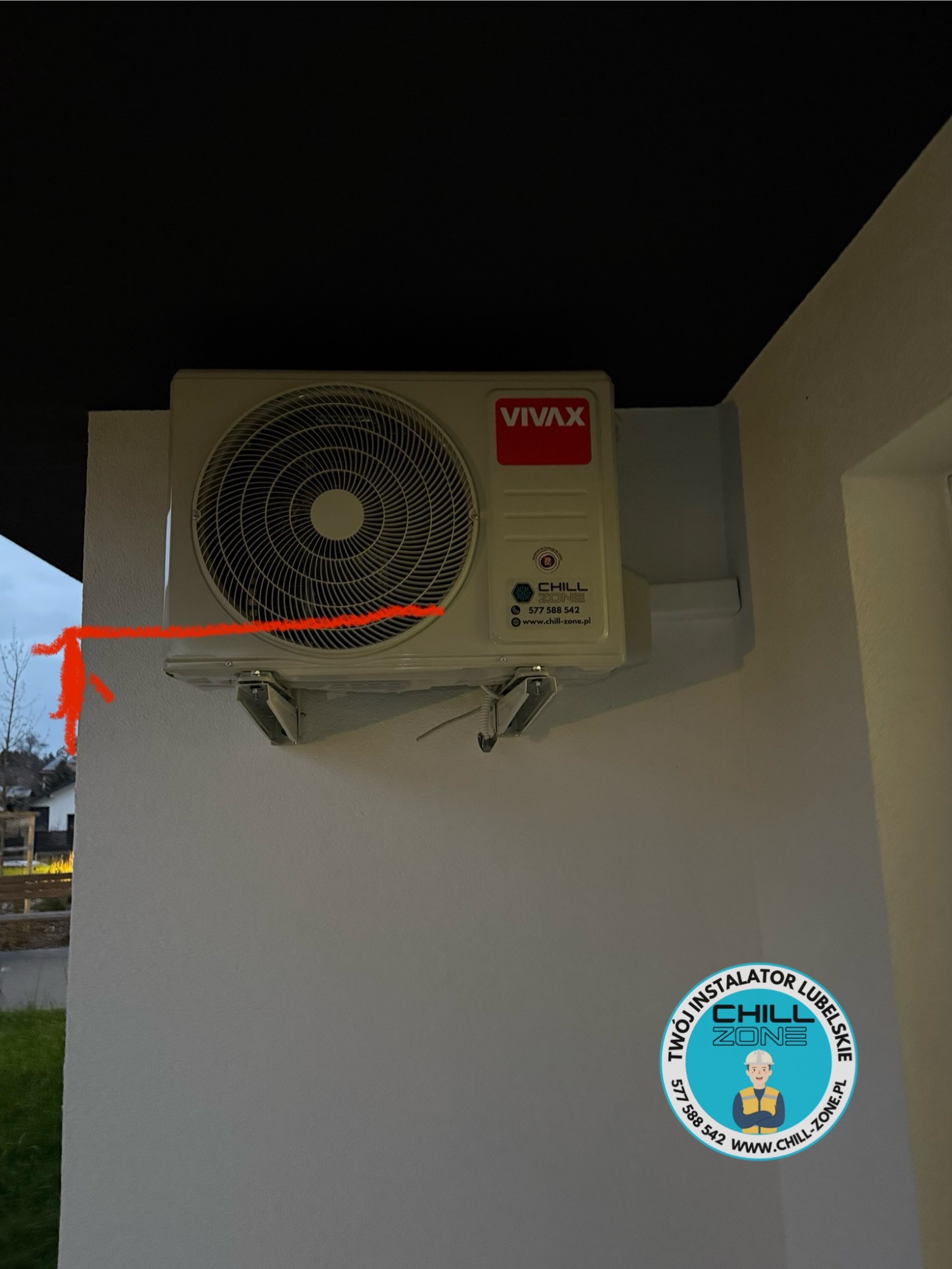 Zamontowana jednostka zewnętrzna klimatyzacji VIVAX na elewacji budynku. Widoczne logo firmy instalacyjnej Chill Zone. Ujęcie z dołu, w tle zieleń i fragment zabudowy.