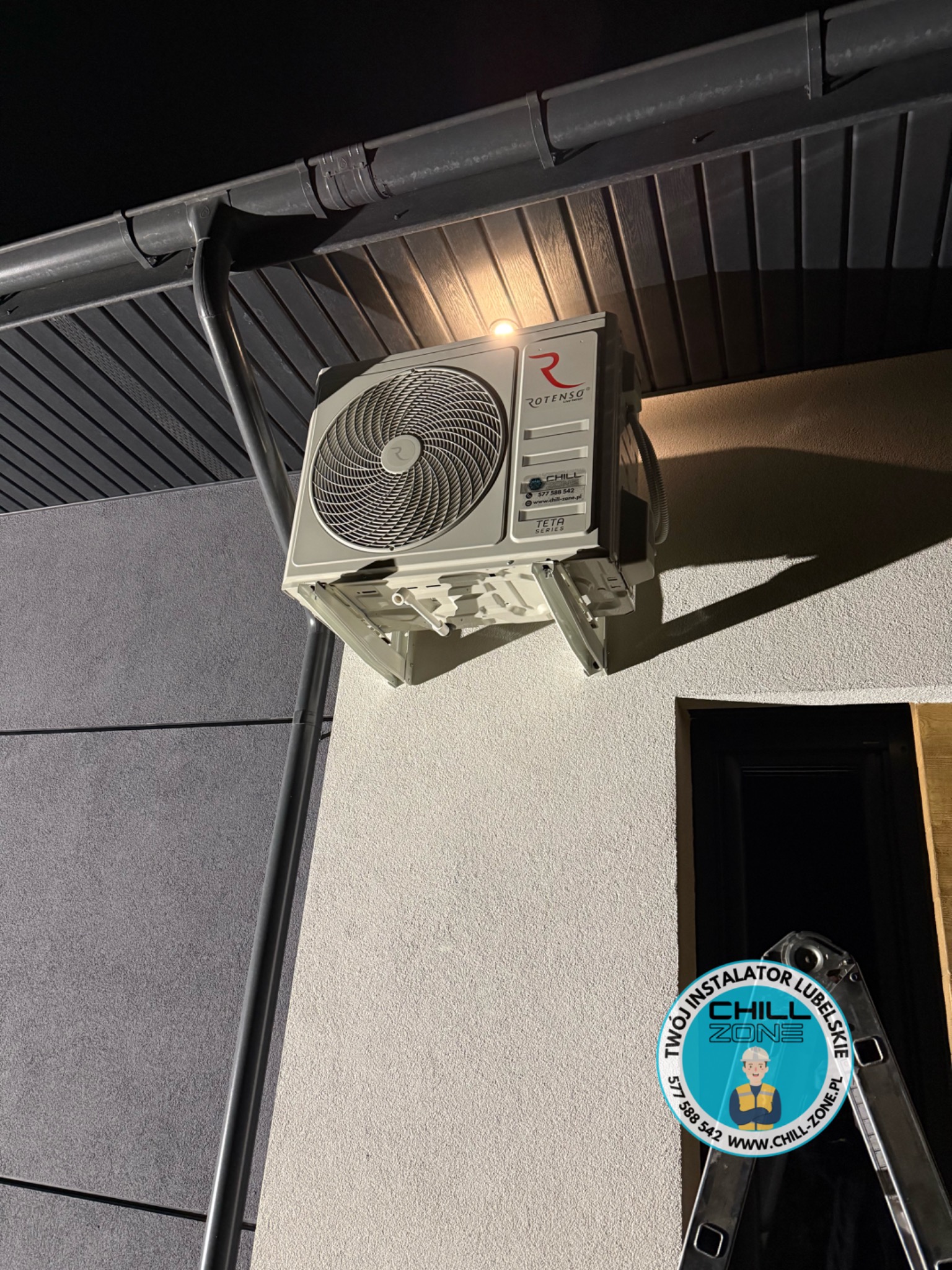 Zamontowana jednostka zewnętrzna klimatyzacji Rotenso Teta Series na elewacji budynku, widoczna drabina i rynna. Oświetlenie nocne.