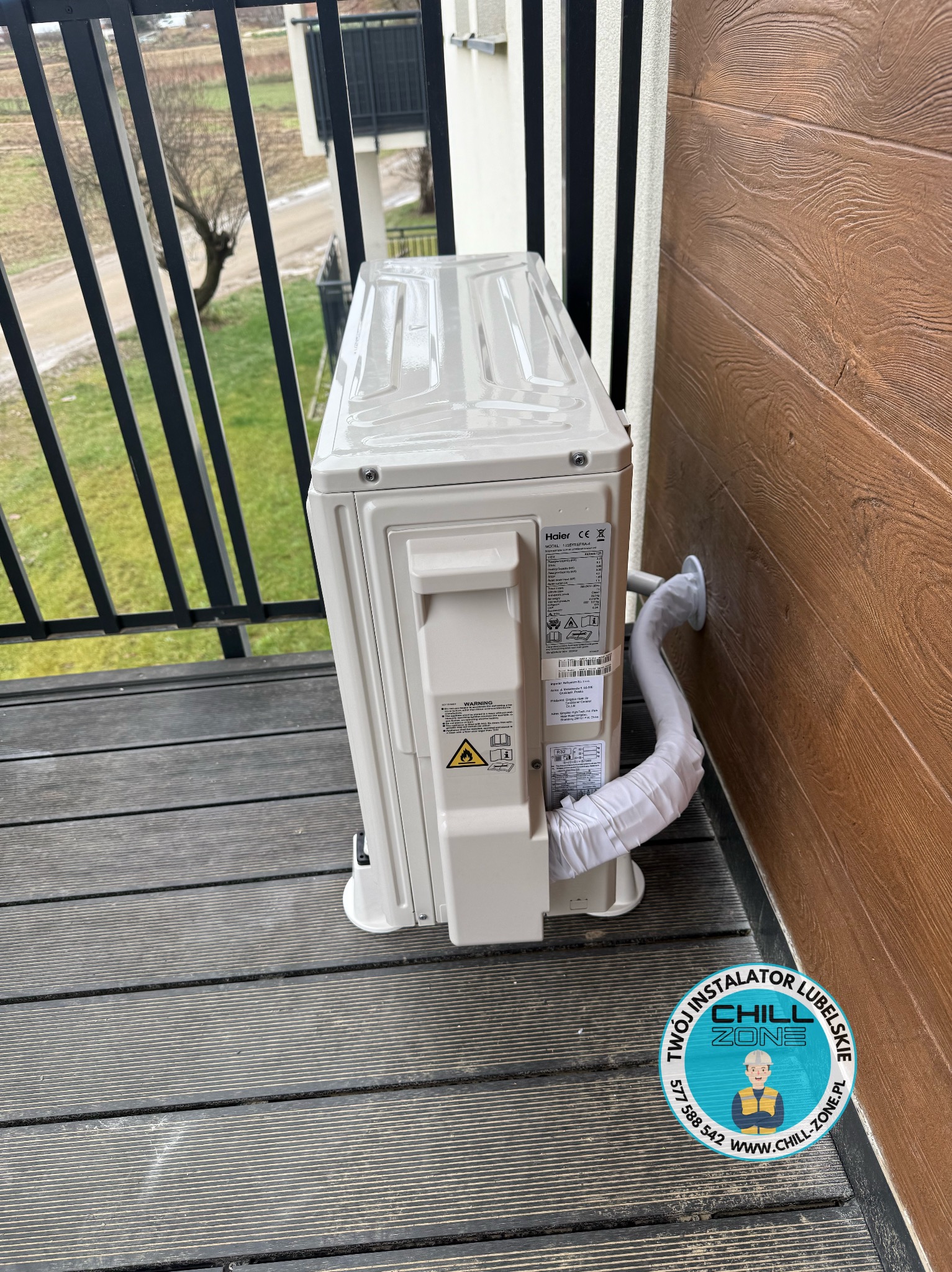 Biała jednostka zewnętrzna klimatyzacji Haier zamontowana na balkonie z drewnianą podłogą i brązową elewacją. Widoczny przewód odprowadzający skropliny.