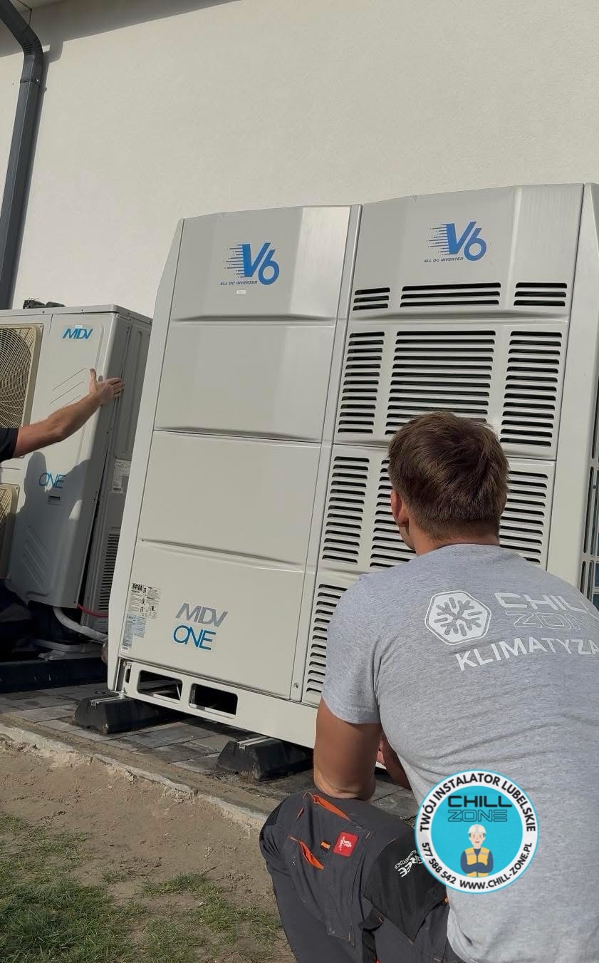 Montaż klimatyzacji MDV ONE V6. Dwóch instalatorów w trakcie pracy przy zewnętrznych jednostkach klimatyzacyjnych na tle jasnej ściany budynku.