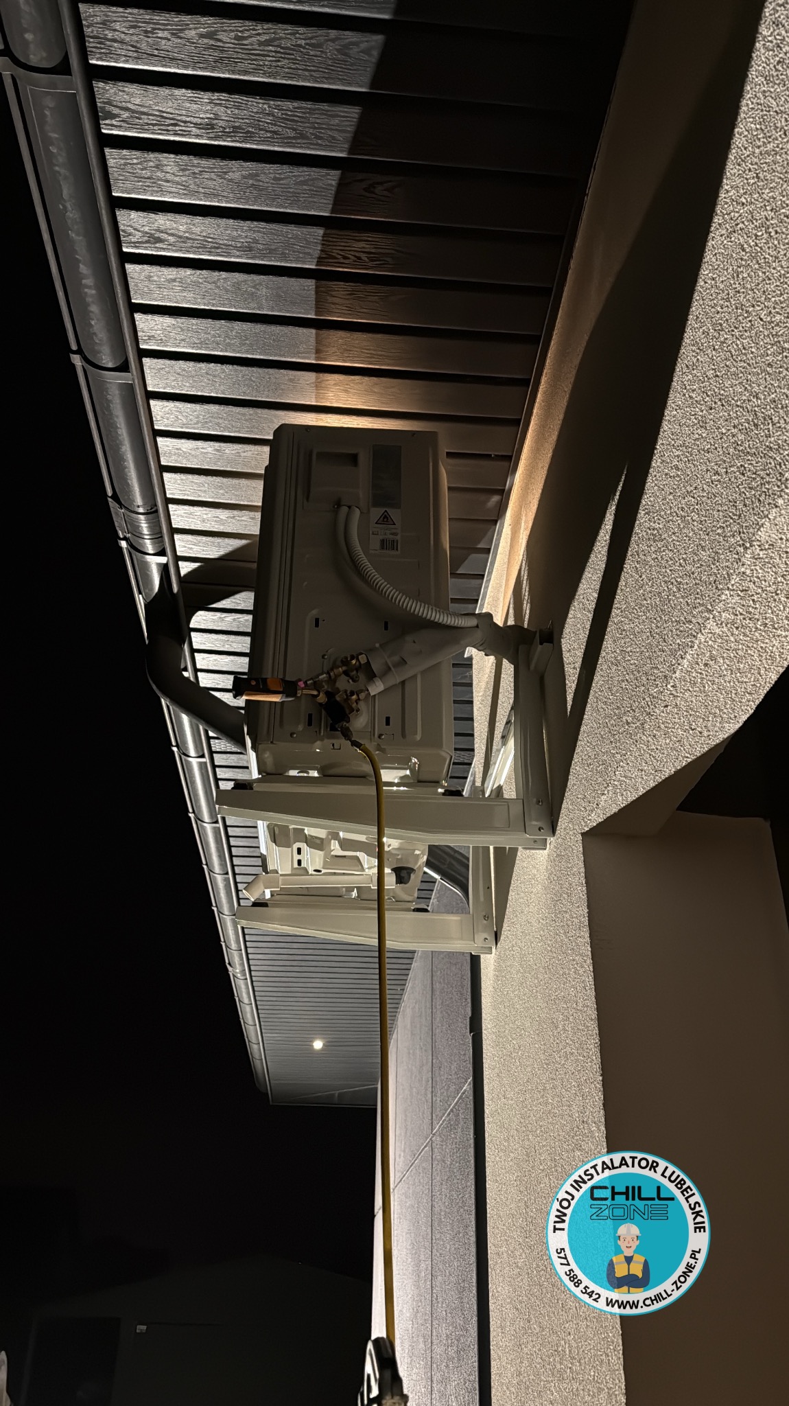 Zewnętrzna jednostka klimatyzacji zamontowana na ścianie budynku, widoczna instalacja i przewody. Oświetlenie nocne podkreśla detale montażu.