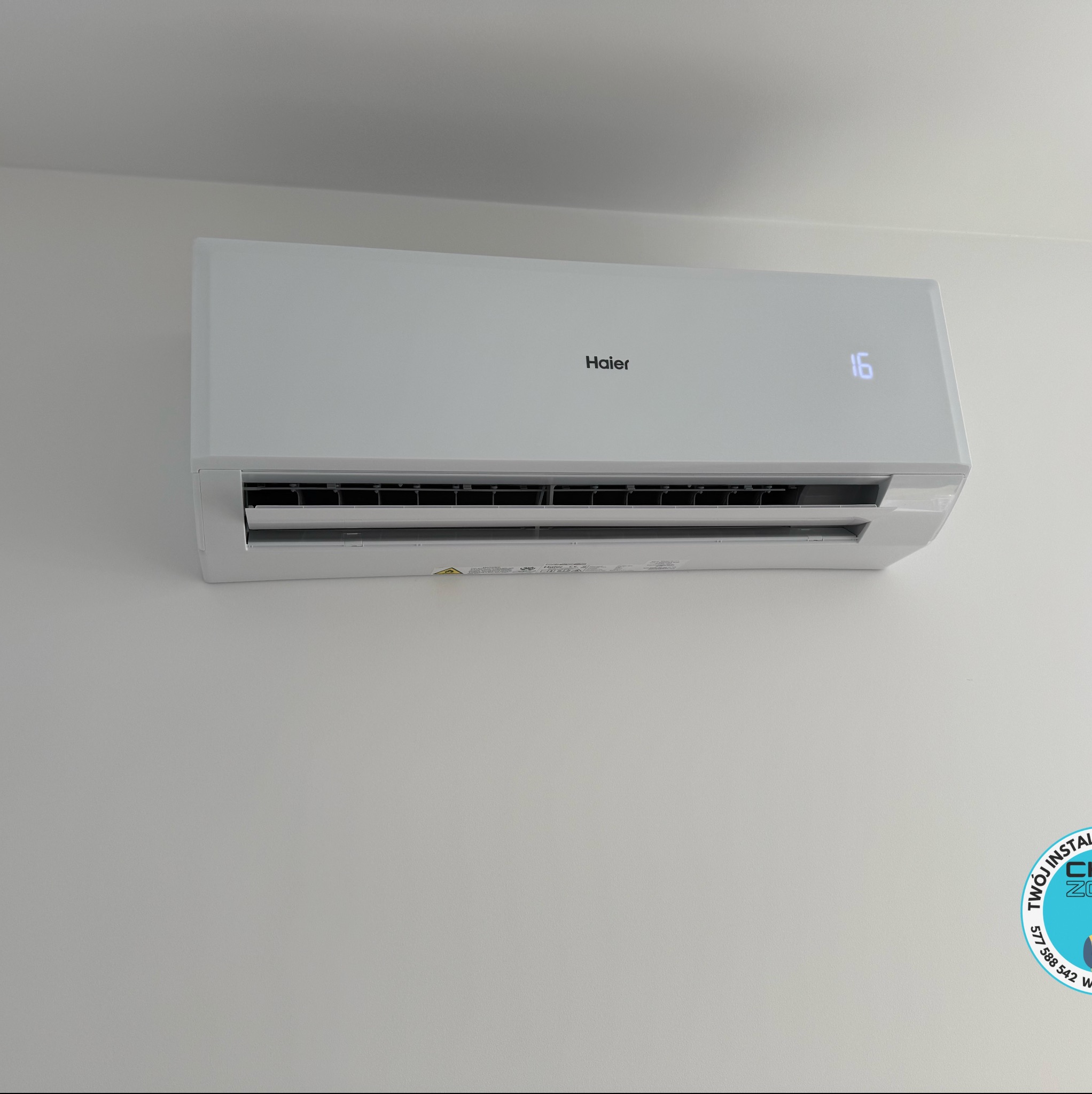 Biały klimatyzator Haier z wyświetlaczem temperatury (16 stopni) zamontowany na jasnej ścianie. Widoczne detale urządzenia i subtelne oświetlenie.