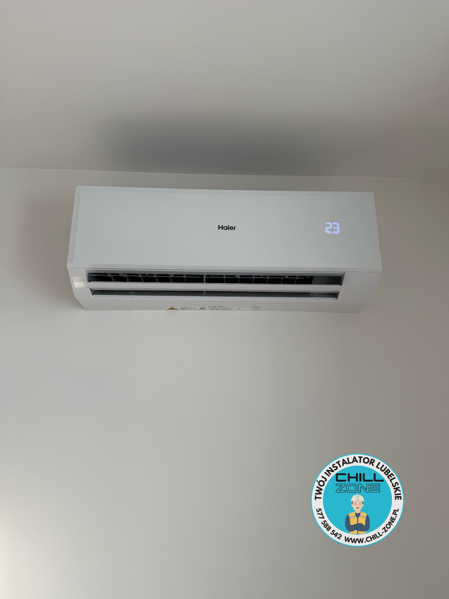 Biały klimatyzator Haier z wyświetlaczem 23°C, zamontowany na jasnej ścianie. W prawym dolnym rogu naklejka z logo firmy instalacyjnej.
