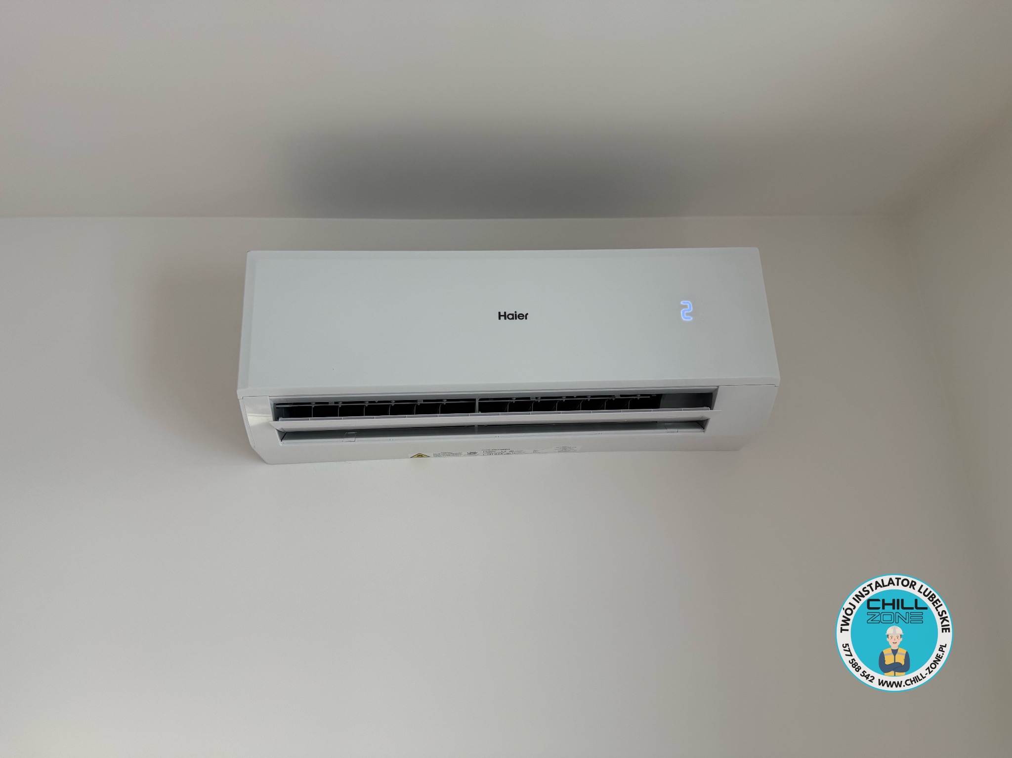 Biały klimatyzator Haier z wyświetlaczem temperatury zamontowany na jasnej ścianie, widoczne logo firmy instalacyjnej w prawym dolnym rogu.