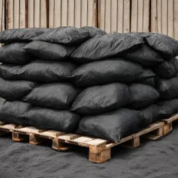 Stos work&oacute;w z czarnym granulatem na drewnianej palecie, przed metalowym ogrodzeniem, z widocznymi śladami pyłu na podłożu.