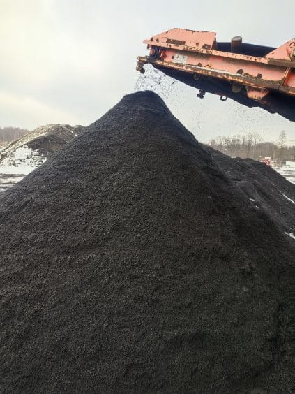 Kopalnia odkrywkowa: hałda ciemnego, drobnego kruszywa, z której wysypuje się materiał z pomarańczowego podajnika taśmowego. W tle widać drzewa i resztki śniegu.