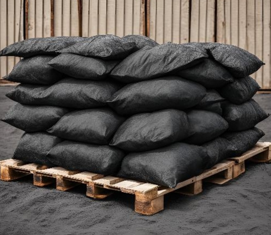 Stos worków z czarnym granulatem na drewnianej palecie, przed metalowym ogrodzeniem, z widocznymi śladami pyłu na podłożu.