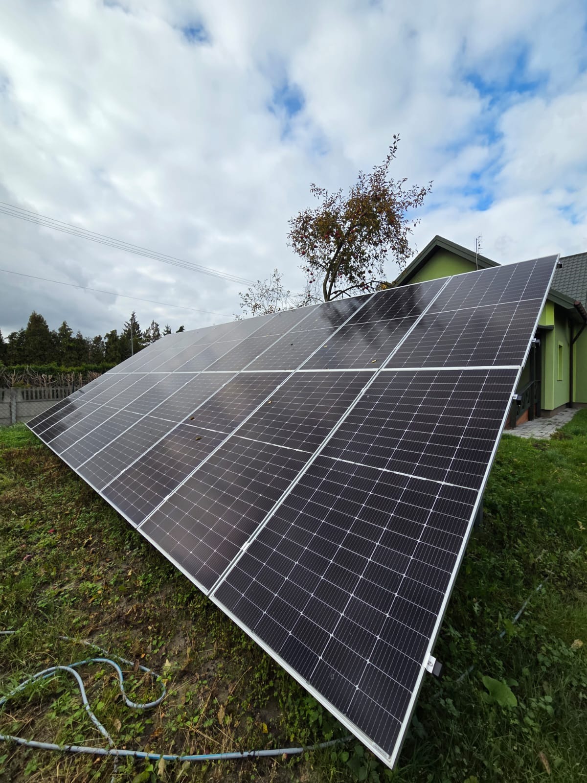 Panel fotowoltaiczny na trawie przed zielonym domem, widok z dołu. Błękitne niebo z chmurami w tle. Instalacja paneli słonecznych na posesji.