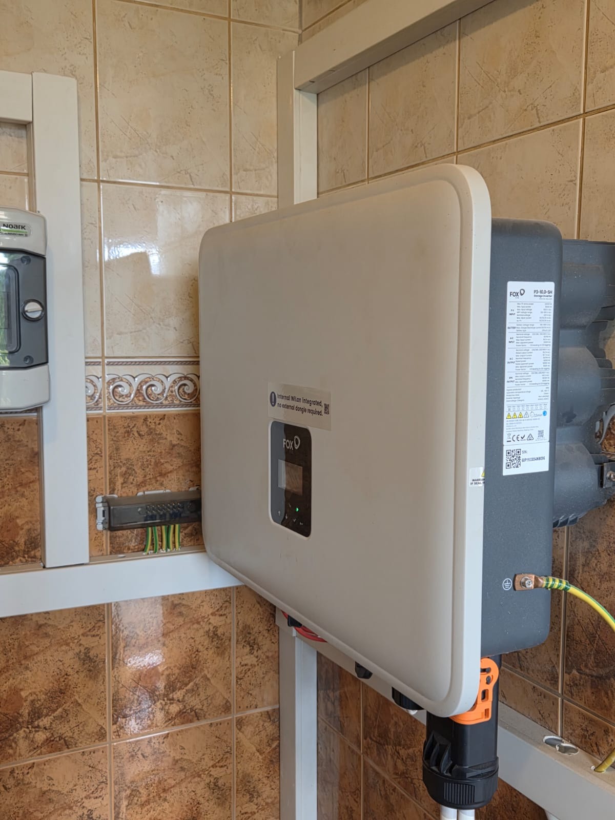 Zamontowany inwerter FOX na ścianie z brązowych płytek. Widoczne przewody i elementy instalacji. Inwerter z wyświetlaczem i naklejką informacyjną.