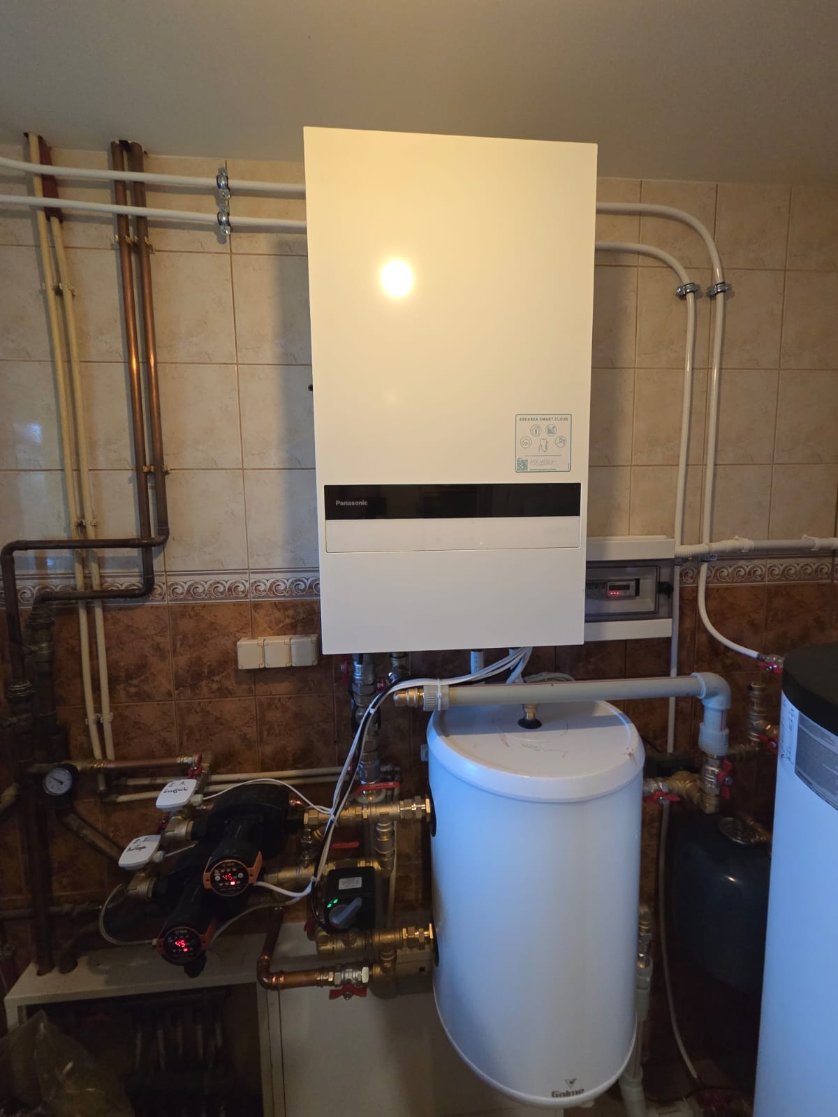 Biały, wiszący piec gazowy Panasonic z podłączonymi rurami miedzianymi i zbiornikiem Galmet w pomieszczeniu z brązowymi płytkami. Widoczne liczniki i zawory.