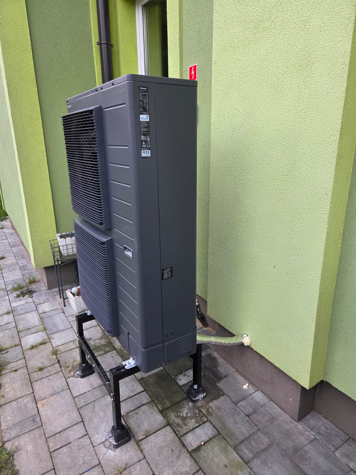 Szara pompa ciepła na czarnym stelażu, zamontowana przy zielonej ścianie budynku. Widoczny fragment instalacji odprowadzającej wodę. Ujęcie z boku, na zewnątrz.