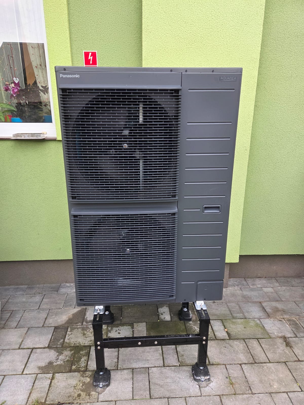 Szara pompa ciepła Panasonic Aquarea na czarnym stojaku, ustawiona na kostce brukowej przed zieloną ścianą budynku z oknem. Widoczny znak ostrzegawczy.