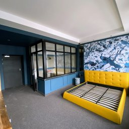 ALEX-BUD PRO Oleksii Filkov - Pokój z niebieskimi ścianami, dekoracyjną tapetą z motywem ptaków i żółtym łóżkiem bez materaca, oddzielony przeszkloną ścianą od łazienki.