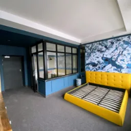 Pokój z niebieskimi ścianami, dekoracyjną tapetą z motywem ptaków i żółtym łóżkiem bez materaca, oddzielony przeszkloną ścianą od łazienki.