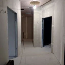 Korytarz w trakcie prac wykończeniowych: ściany pokryte płytami kartonowo-gipsowymi, widoczne łączenia do szpachlowania, sufit z okrągłym otworem na oświetlenie, grzejnik żeliwny w rogu.