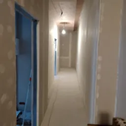 Korytarz w trakcie prac wykończeniowych, ściany pokryte gładzią z widocznymi łatami, sufit z elementem dekoracyjnym w kolorze terakoty, widoczne kable oświetleniowe, w tle otwarte drzwi...