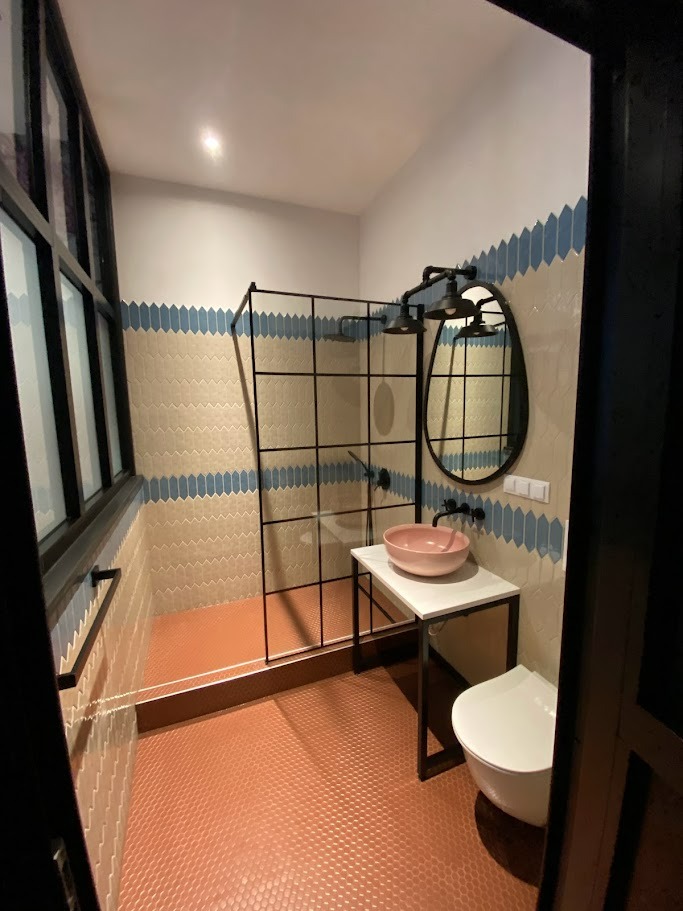 Nowoczesna łazienka z kabiną prysznicową typu walk-in z czarną ramą, umywalką w kolorze pudrowego różu na białym blacie z czarną ramą, okrągłym lustrem z czarną ramą i terakotową podłogą z drobnymi...