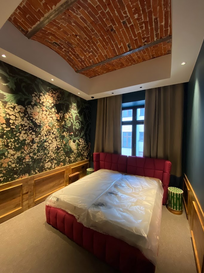 Sypialnia po remoncie z łóżkiem przykrytym folią ochronną, dekoracyjna tapeta w kwiaty i sufit z ceglanym sklepieniem łukowym.