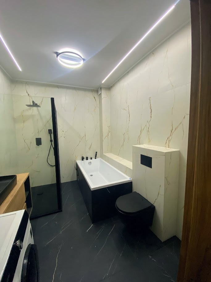 Nowoczesna łazienka z czarnymi akcentami: kabina prysznicowa typu walk-in, wanna w obudowie imitującej czarny marmur, wisząca toaleta i płytki ścienne w kolorze złamanej bieli z marmurkowym wzorem,...