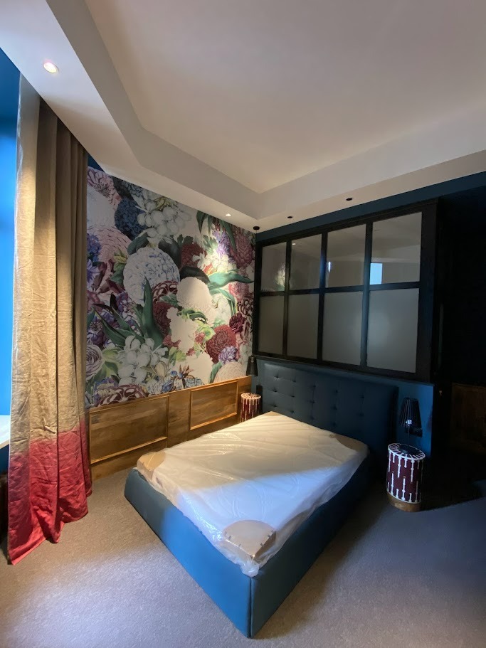 Sypialnia po remoncie z tapetą w kwiaty na ścianie, drewnianą boazerią i niebieskim łóżkiem, okno w stylu industrialnym z czarnymi szprosami.