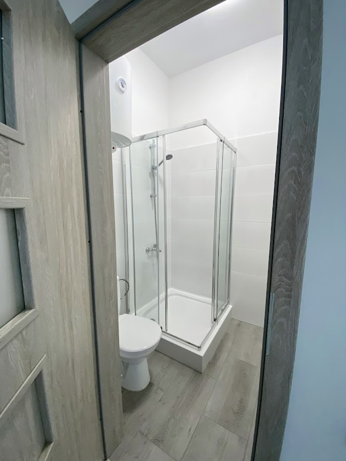 Nowoczesna łazienka z kabiną prysznicową z chromowanymi profilami, białą ceramiką sanitarną i grzejnikiem elektrycznym nad toaletą, widoczna zza otwartych drzwi o teksturze jasnego drewna.