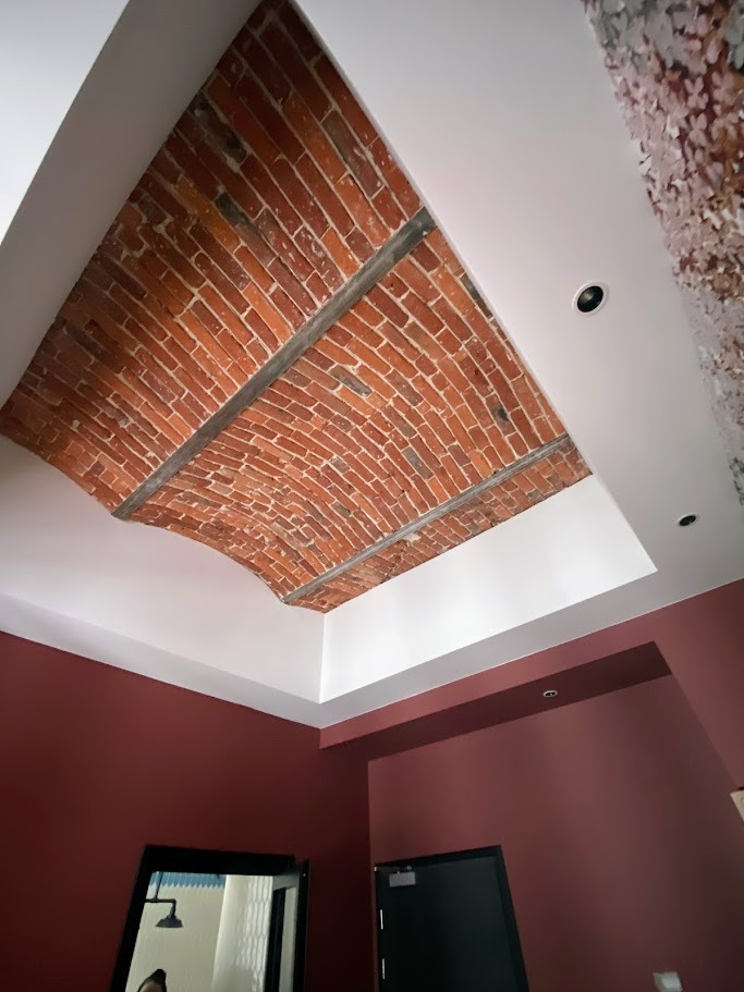 Ceglany łuk na suficie podtrzymywany przez metalowe belki, otoczony białym obramowaniem i oświetleniem punktowym, ściany w kolorze burgundowym, widoczne drzwi i lustro.